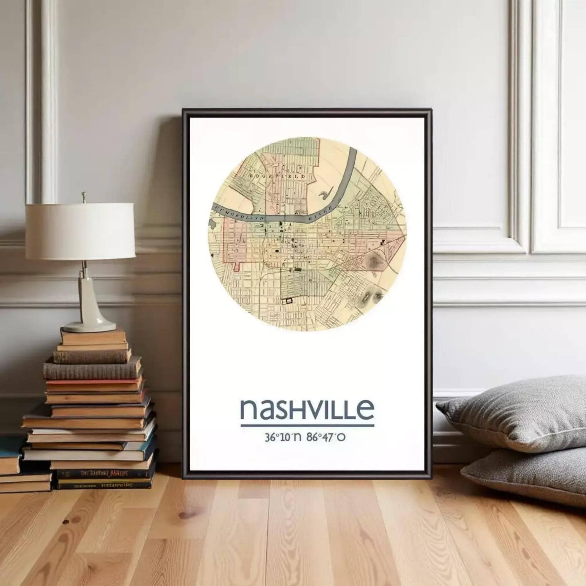 A framed map of Nashville, Tennessee, with the text " Nashville 36°10'11" 86°47'7"", placed on a wooden floor next to a stack of books and a lamp.