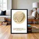 A framed map of Cambridge, England, with the text "Cambridge.ma" and the coordinates "42.22 N 71.96 W" displayed on it, placed on a wooden floor in front of a bookshelf.