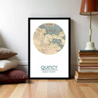A framed map of QuINCY, a coastal city, is displayed on a wall next to a stack of books and a lamp.