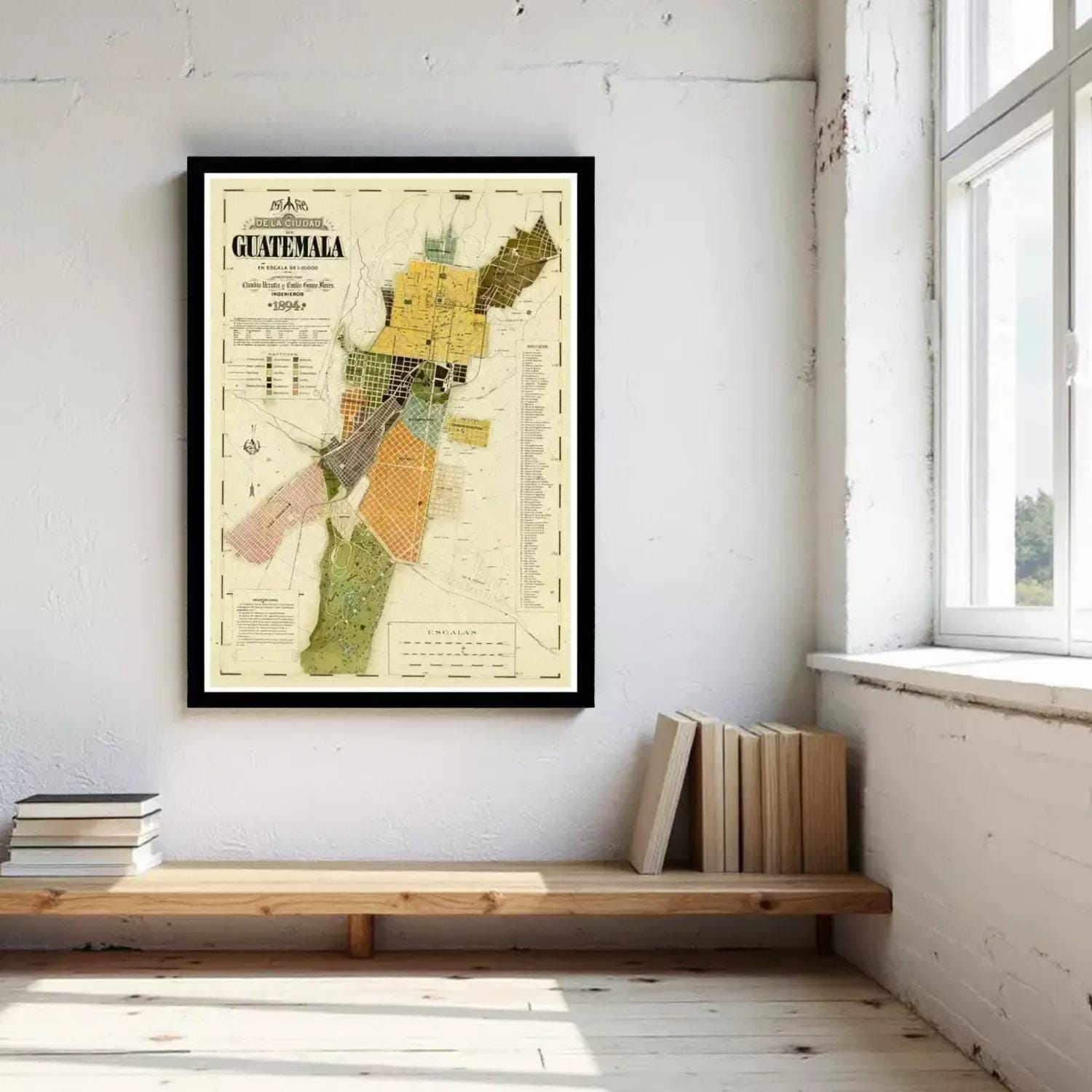 a framed map of Guatemala hanging on a white wall, with a wooden bench and a stack of books placed nearby.