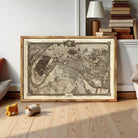A framed map of Paris, France, displayed on a wooden floor in a room with a lamp and bookshelf in the background.
